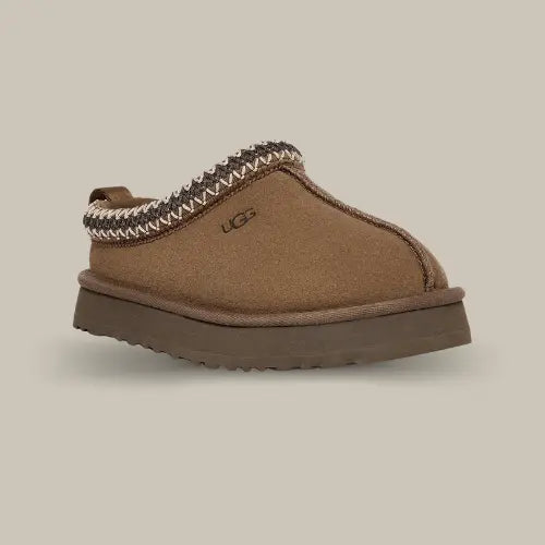 La UGG Tazz Slipper Hickory avec une tige en daim, une tresse marron et blanche en polyester recyclé et sa semelle compensée en EVA de 3.8cm.