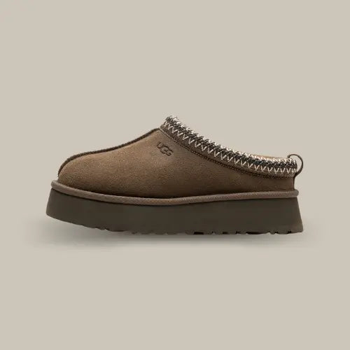 La UGG Tazz Slipper Hickory avec une tige en daim, une tresse marron et blanche en polyester recyclé et sa semelle compensée en EVA de 3.8cm.